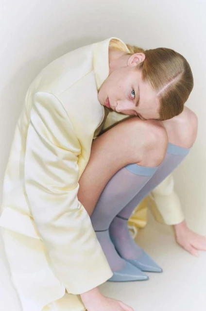 An adult woman in a crouched pose wearing a light yellow jacket, hosiery, and matching shoes in a bright studio setting.