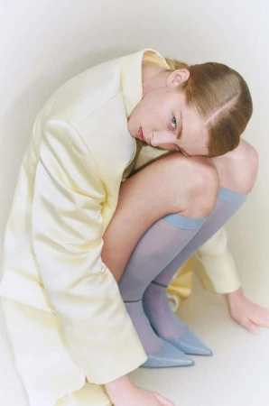 An adult woman in a crouched pose wearing a light yellow jacket, hosiery, and matching shoes in a bright studio setting.