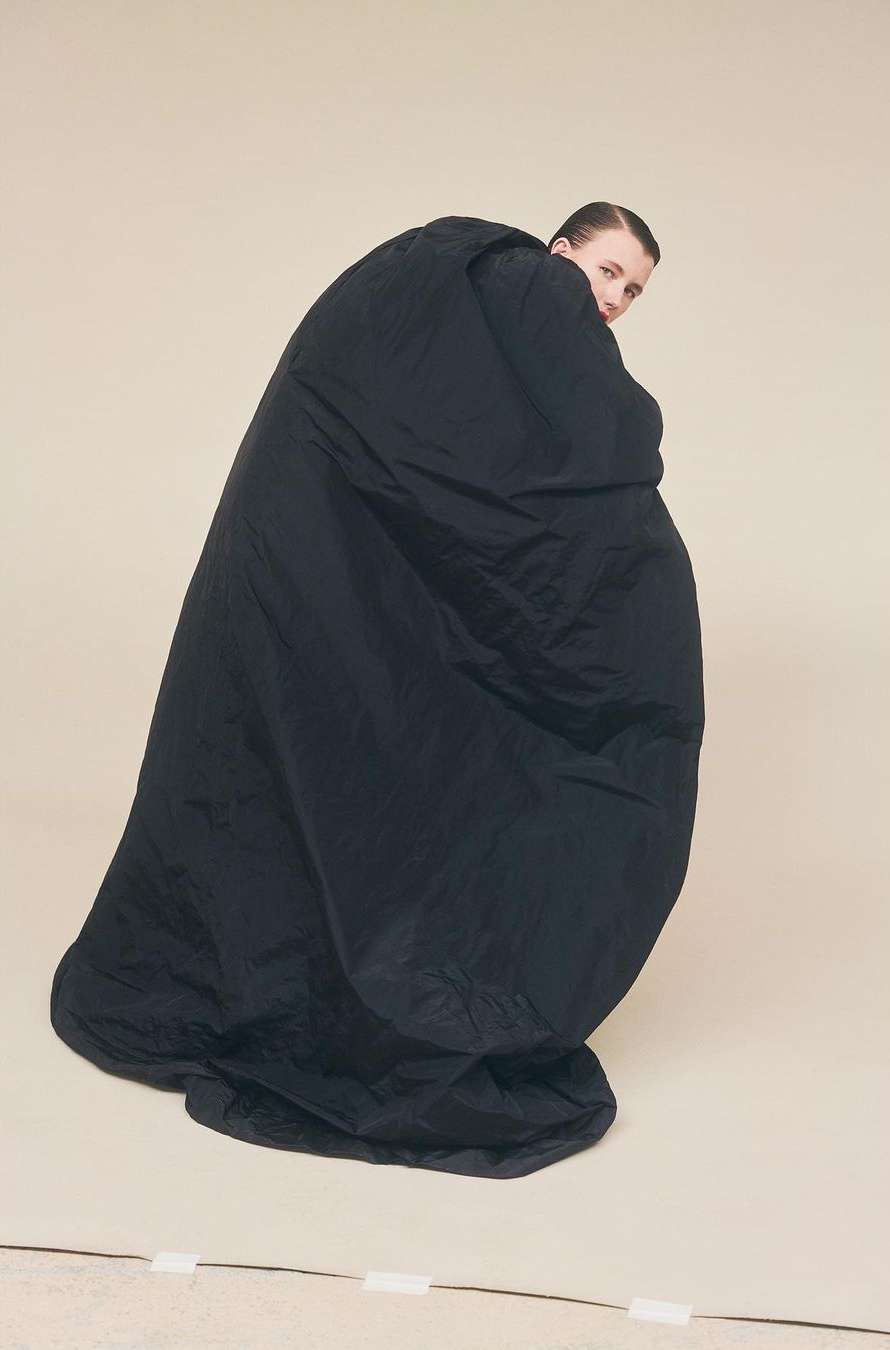 Woman in black oversized garment poses in a studio. Fashion editorial portrait features a model looking over her shoulder in a beige setting.