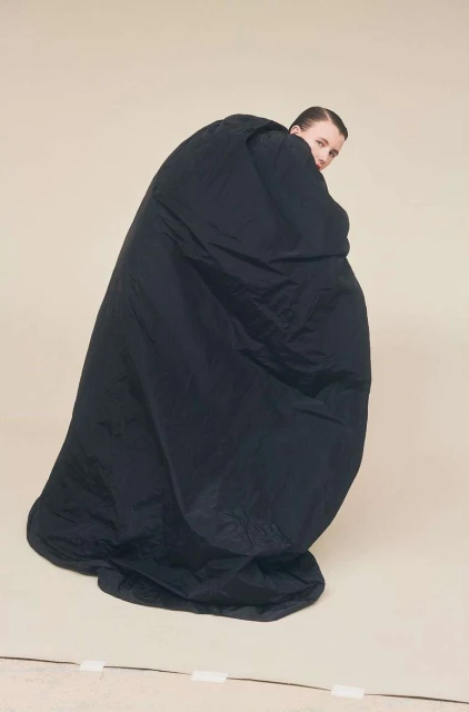 Woman in black oversized garment poses in a studio. Fashion editorial portrait features a model looking over her shoulder in a beige setting.