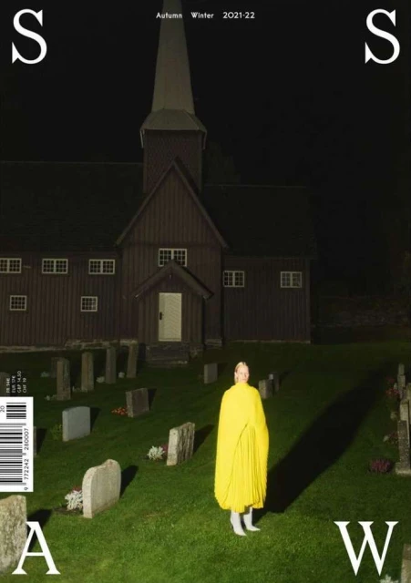 Fashion editorial featuring a model in a yellow cape, standing in a cemetery with a church in the background at nighttime.