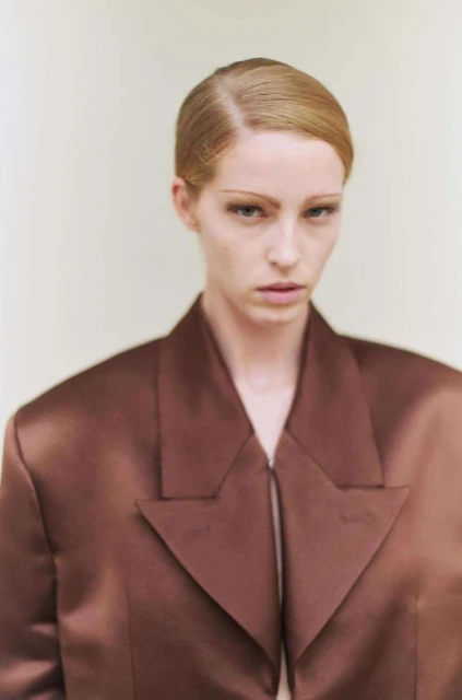 Portrait of an adult woman with light blonde hair wearing a brown satin blazer against an off-white background in a studio environment.
