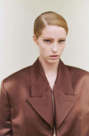 Portrait of a blonde woman wearing a structured brown satin jacket against a plain light background by Ola Rindal.