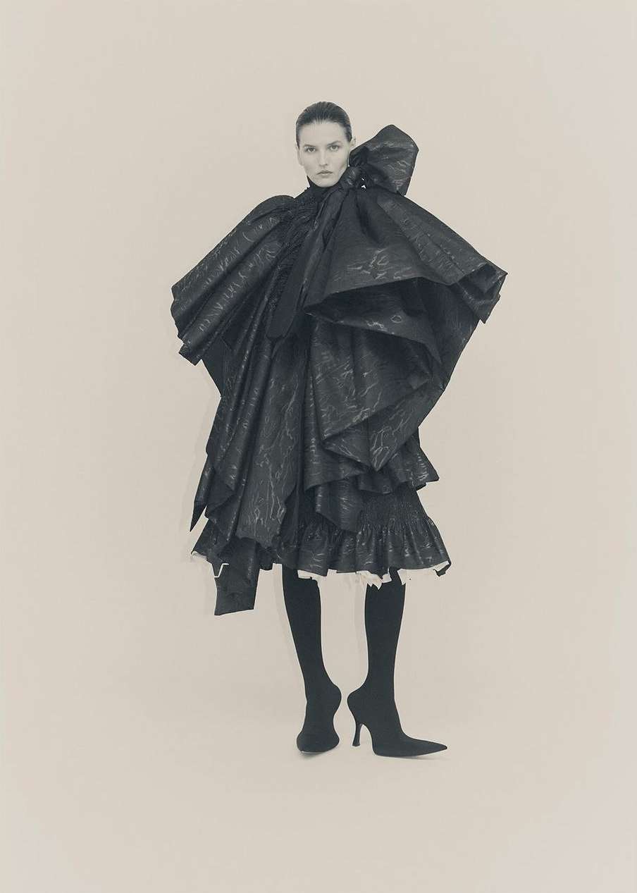 Model in voluminous black sculptural dress with large bow shoulder detail and dark tights against neutral background