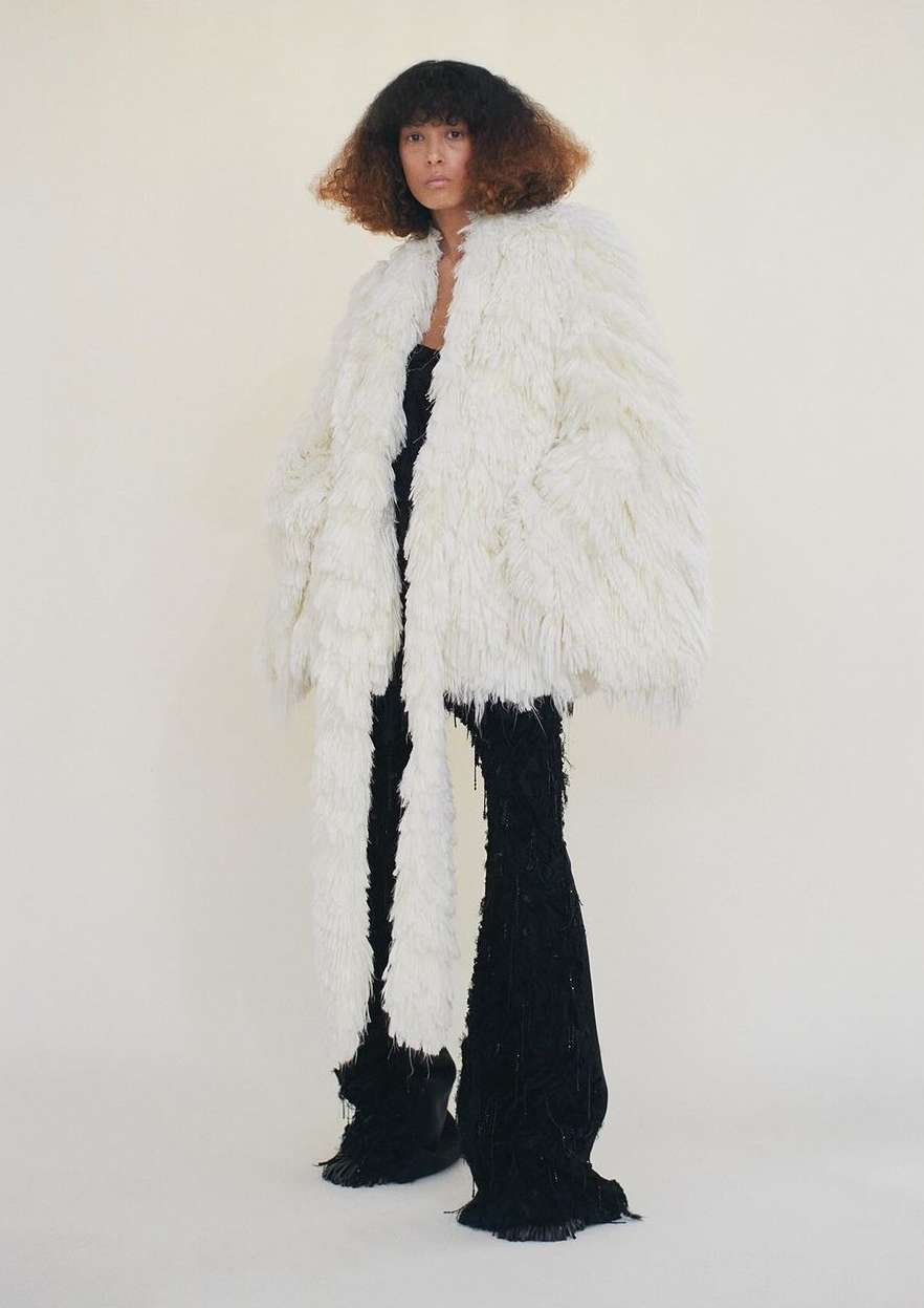 A model with curly hair poses in an off-white shaggy coat and black knit pants against a light neutral background in a full length studio shot.