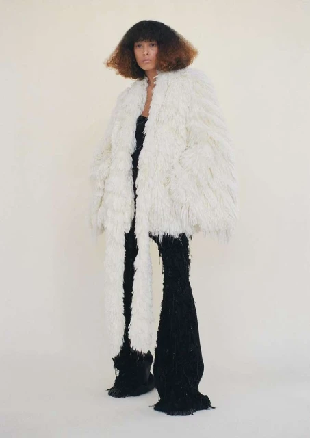 A model with curly hair poses in an off-white shaggy coat and black knit pants against a light neutral background in a full length studio shot.