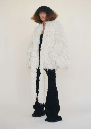 A model with curly hair poses in an off-white shaggy coat and black knit pants against a light neutral background in a full length studio shot.