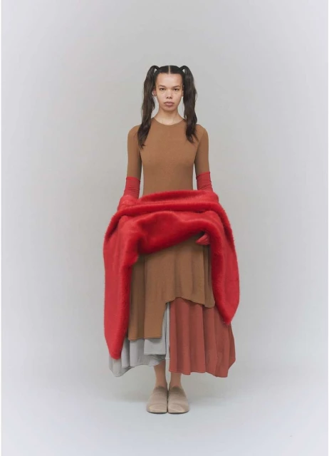 Full-length portrait of a young woman with braided pigtails wearing a brown dress and holding a large red textured scarf.