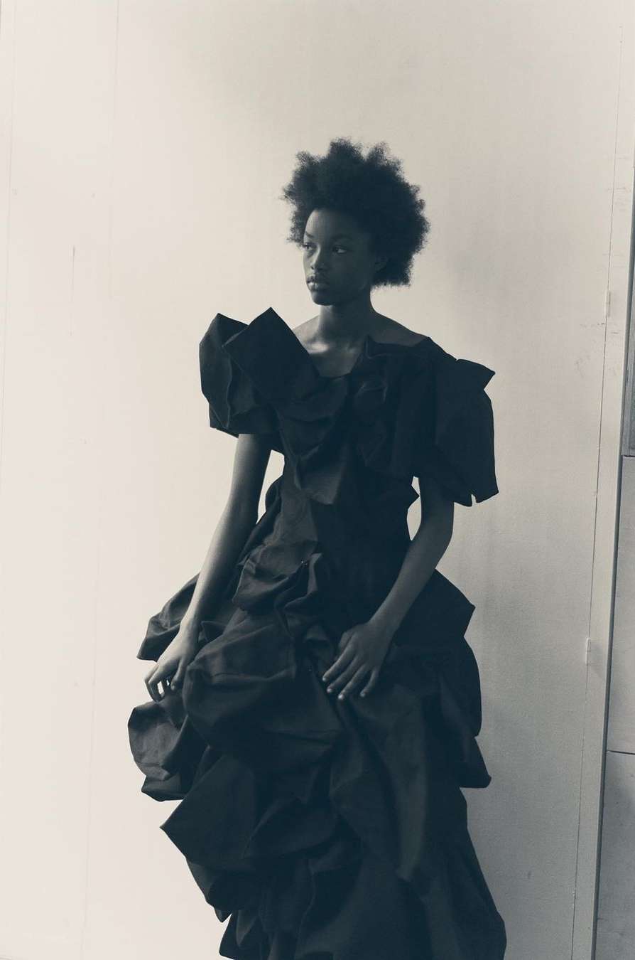 Black woman wearing a sculptural black dress with voluminous ruffles against a plain studio background by Ola Rindal.
