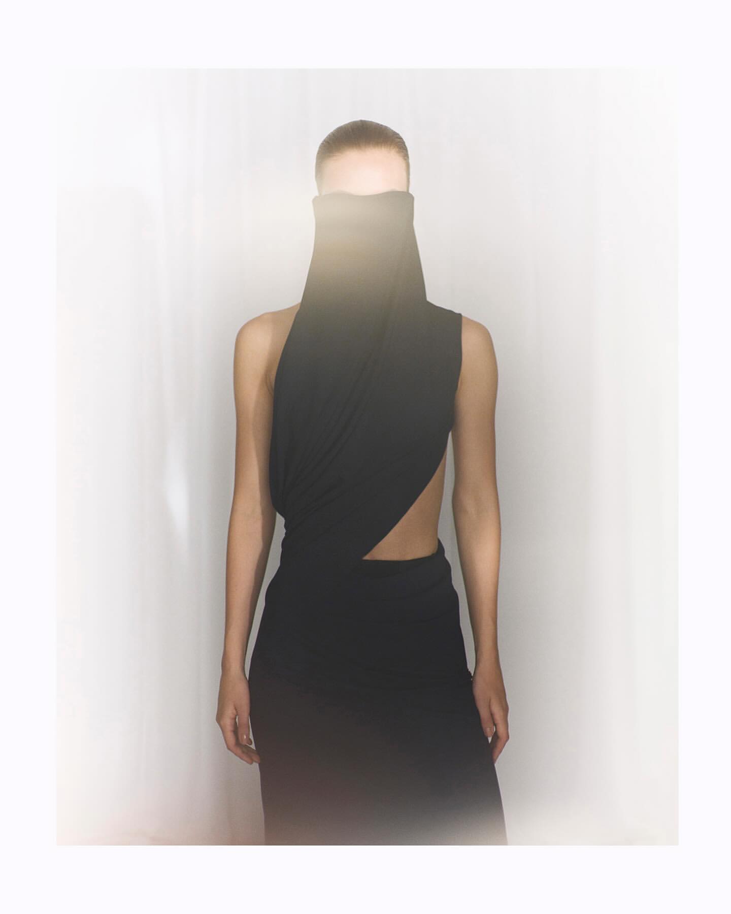Model wearing a sculptural black high-neck draped garment with a bright lens flare obscuring the upper face against a white backdrop.