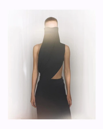 Model wearing a sculptural black high-neck draped garment with a bright lens flare obscuring the upper face against a white backdrop.