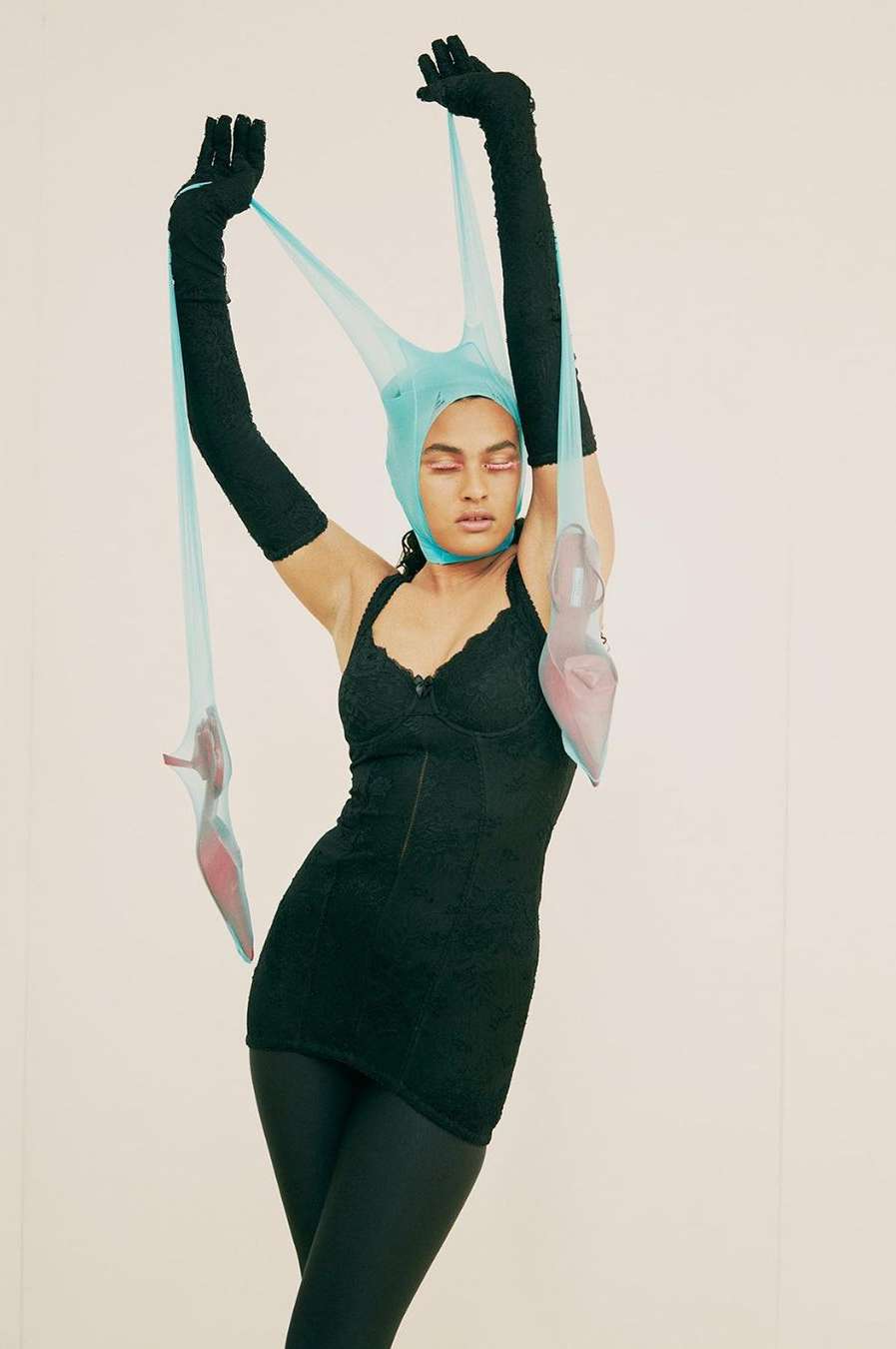 Woman in black dress and headdress poses with suspended transparent heels against a pale backdrop, fashion-forward presentation.
