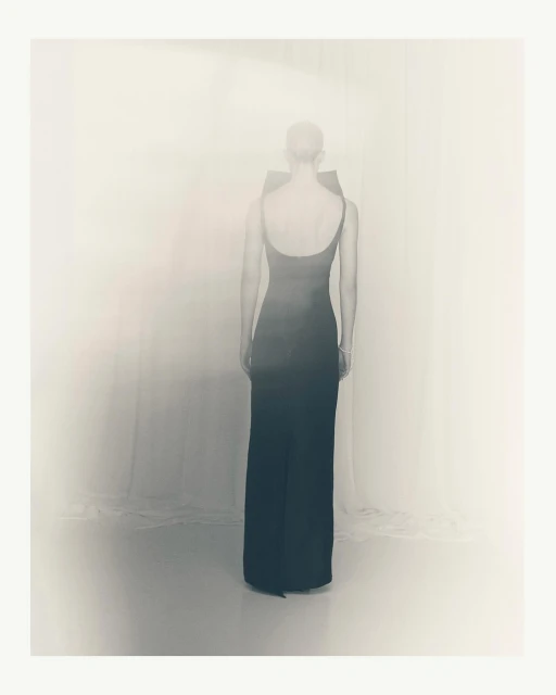 Back view of an adult woman wearing a long black dress with straps, standing in an indoor setting with soft, diffused lighting.