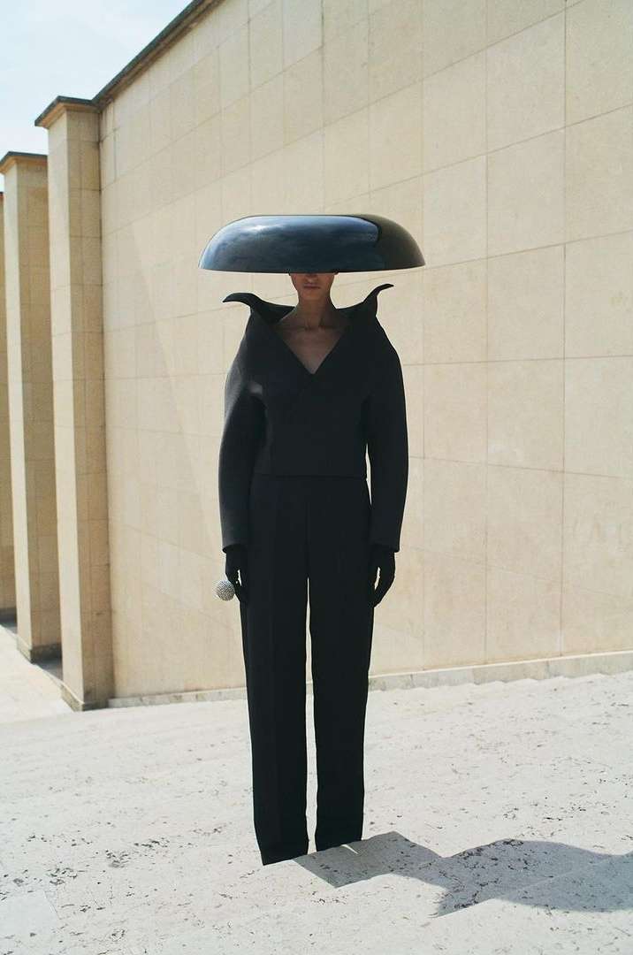 Woman in stylish black outfit and large hat stands on a stone-paved surface, posing in front of a light-beige building with columns under bright daylight.