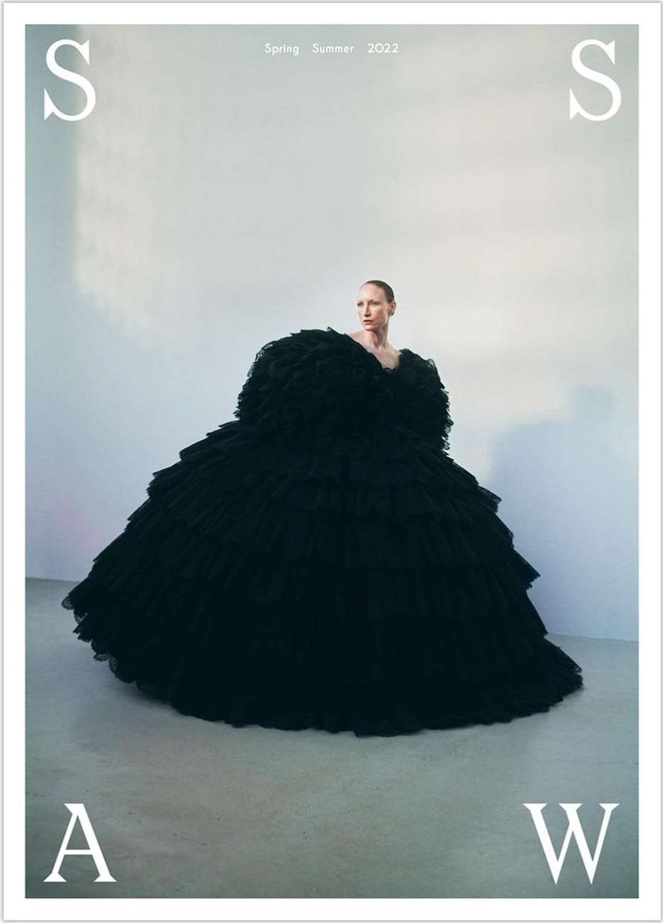 Fashion editorial shot of a female model in a large black ruffled dress against a light grey background, styled as a magazine cover.