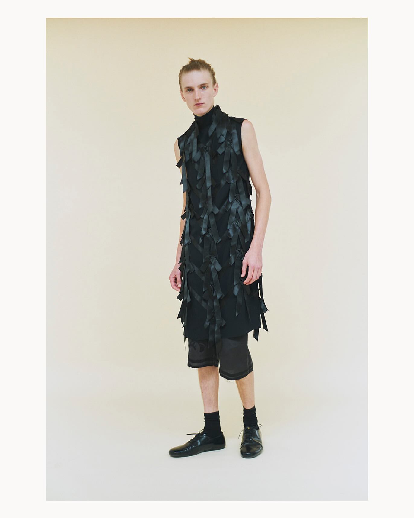 Male model poses in a black, avant-garde sleeveless top with dark shorts and shoes against a neutral background in a studio setting.