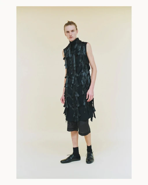 Male model poses in a black, avant-garde sleeveless top with dark shorts and shoes against a neutral background in a studio setting.