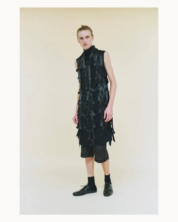 Male model poses in a black, avant-garde sleeveless top with dark shorts and shoes against a neutral background in a studio setting.