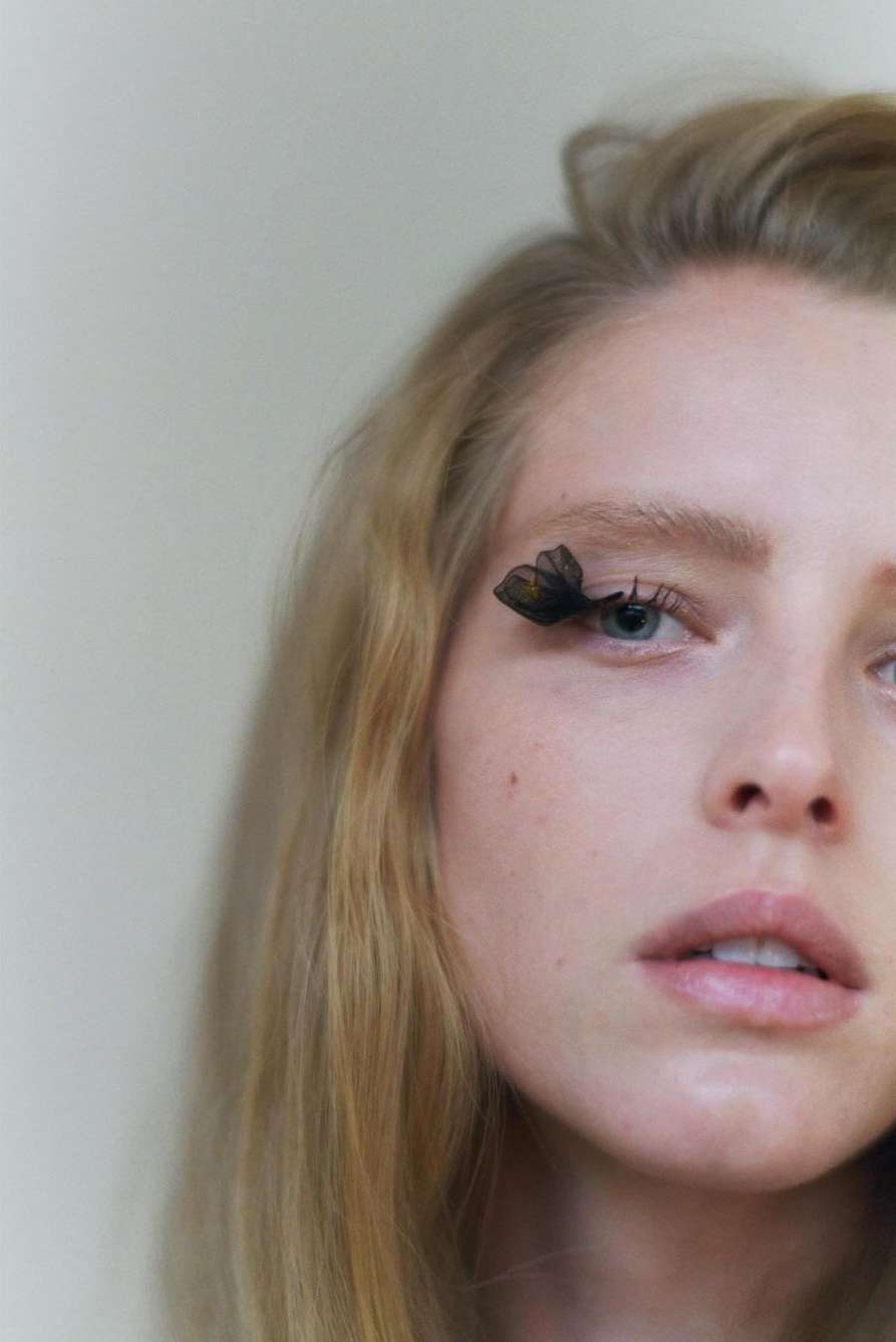 Close-up portrait of a woman with blond hair and blue eyes wearing butterfly-shaped false eyelashes against a soft, neutral background.