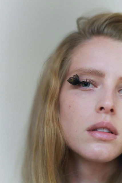 Close-up portrait of a woman with blond hair and blue eyes wearing butterfly-shaped false eyelashes against a soft, neutral background.