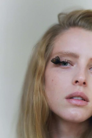 Close-up portrait of a woman with blond hair and blue eyes wearing butterfly-shaped false eyelashes against a soft, neutral background.
