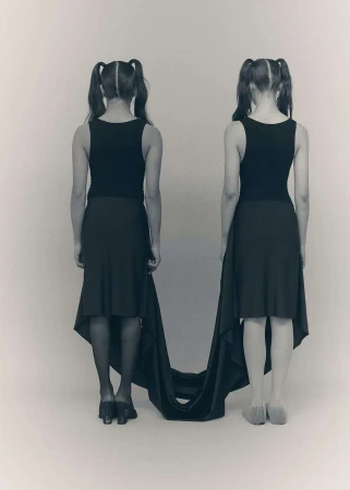 Two young girls, back to the viewer, with ponytails, wear matching black tops and skirts with a long black train.