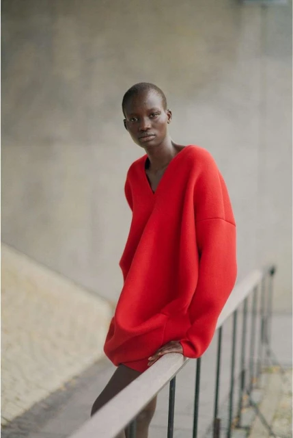 An adult woman poses in a red, oversized sweater in an outdoor fashion shoot against a concrete and metal railing backdrop.