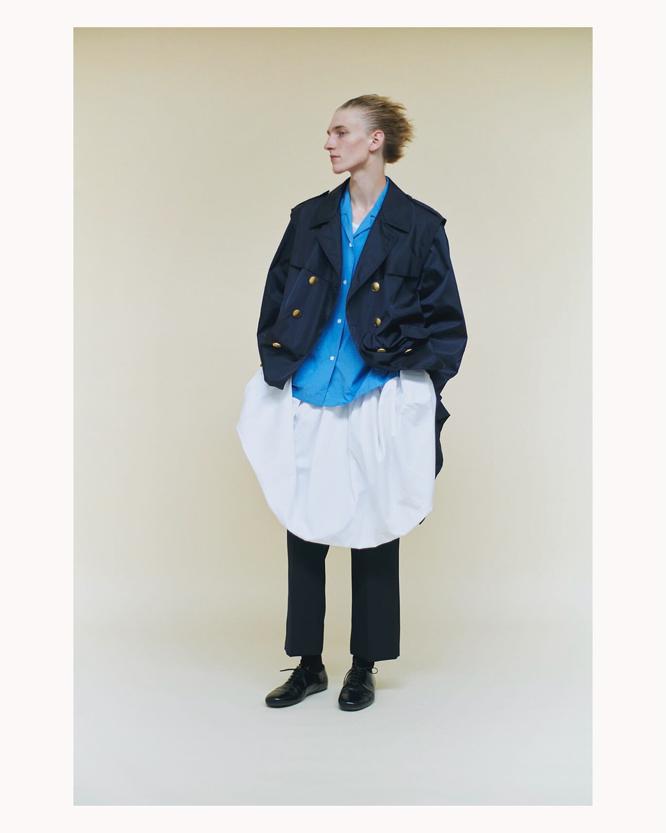 Fashion portrait of a young adult male model in a navy blazer, light blue button-down shirt, white skirt-like garment, and black shoes.