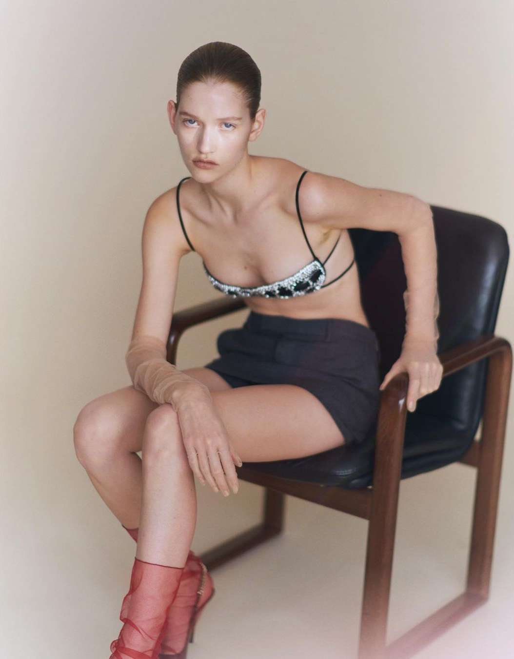 A woman with slicked-back hair poses in a studded bralette, shorts, and red heels while seated on a chair in a minimalist studio.