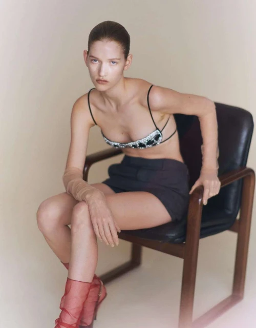 A woman with slicked-back hair poses in a studded bralette, shorts, and red heels while seated on a chair in a minimalist studio.