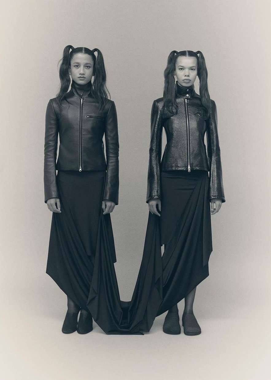 Two young women with pigtails, wearing matching leather jackets and black skirts, pose in a fashion studio, full shot.