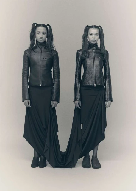 Two young women with pigtails, wearing matching leather jackets and black skirts, pose in a fashion studio, full shot.