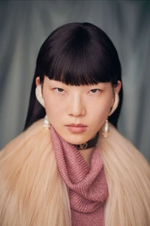 Close-up portrait of a model with dark hair and fringe wearing pearl ear cuffs and a pink textured garment by Ola Rindal.
