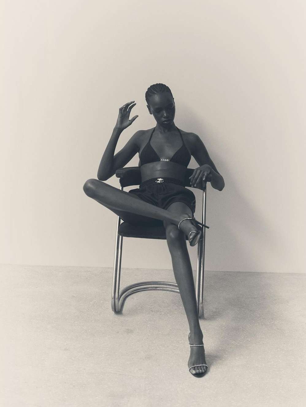 Elegant Black female fashion model sits on chair in bra top and shorts with high-heeled shoes in a minimalist photo studio setting.