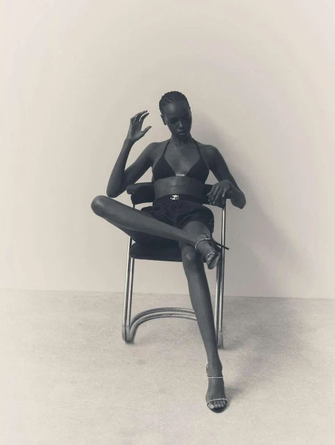 Elegant Black female fashion model sits on chair in bra top and shorts with high-heeled shoes in a minimalist photo studio setting.