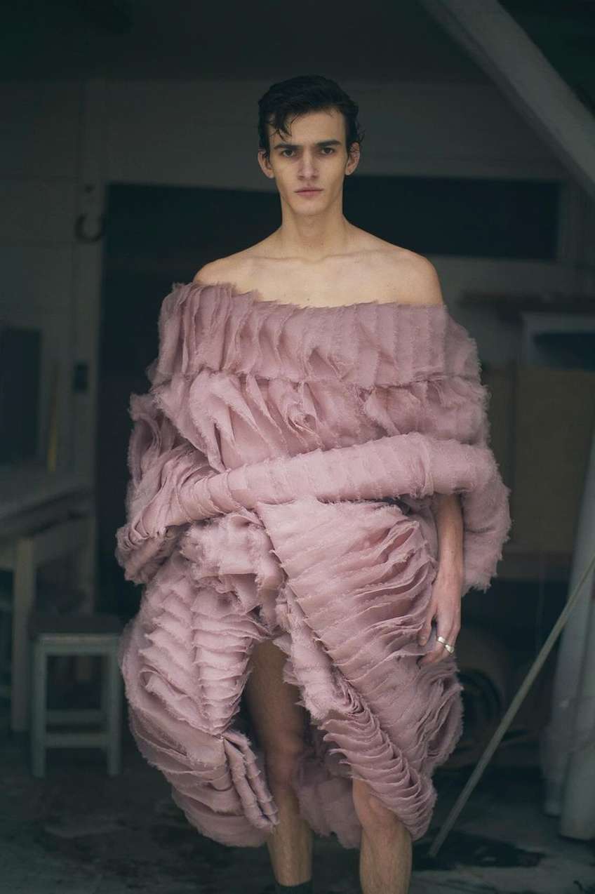 A slender adult male stands in a pink ruffled garment inside a room, the focal point of a fashion editorial style photograph.