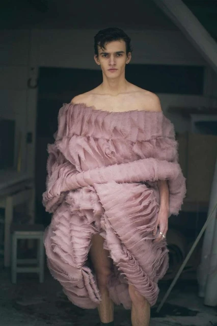 A slender adult male stands in a pink ruffled garment inside a room, the focal point of a fashion editorial style photograph.