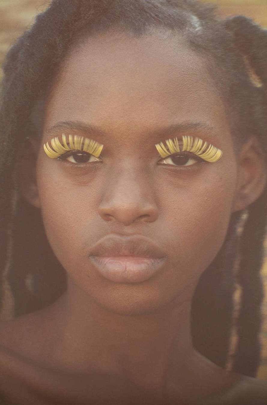 Close-up portrait of a young African woman with elaborate yellow eyelash extensions against a soft yellow and brown background.