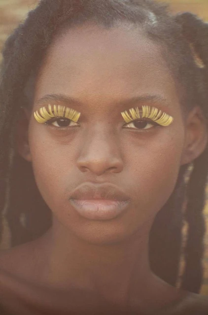 Close-up portrait of a young African woman with elaborate yellow eyelash extensions against a soft yellow and brown background.