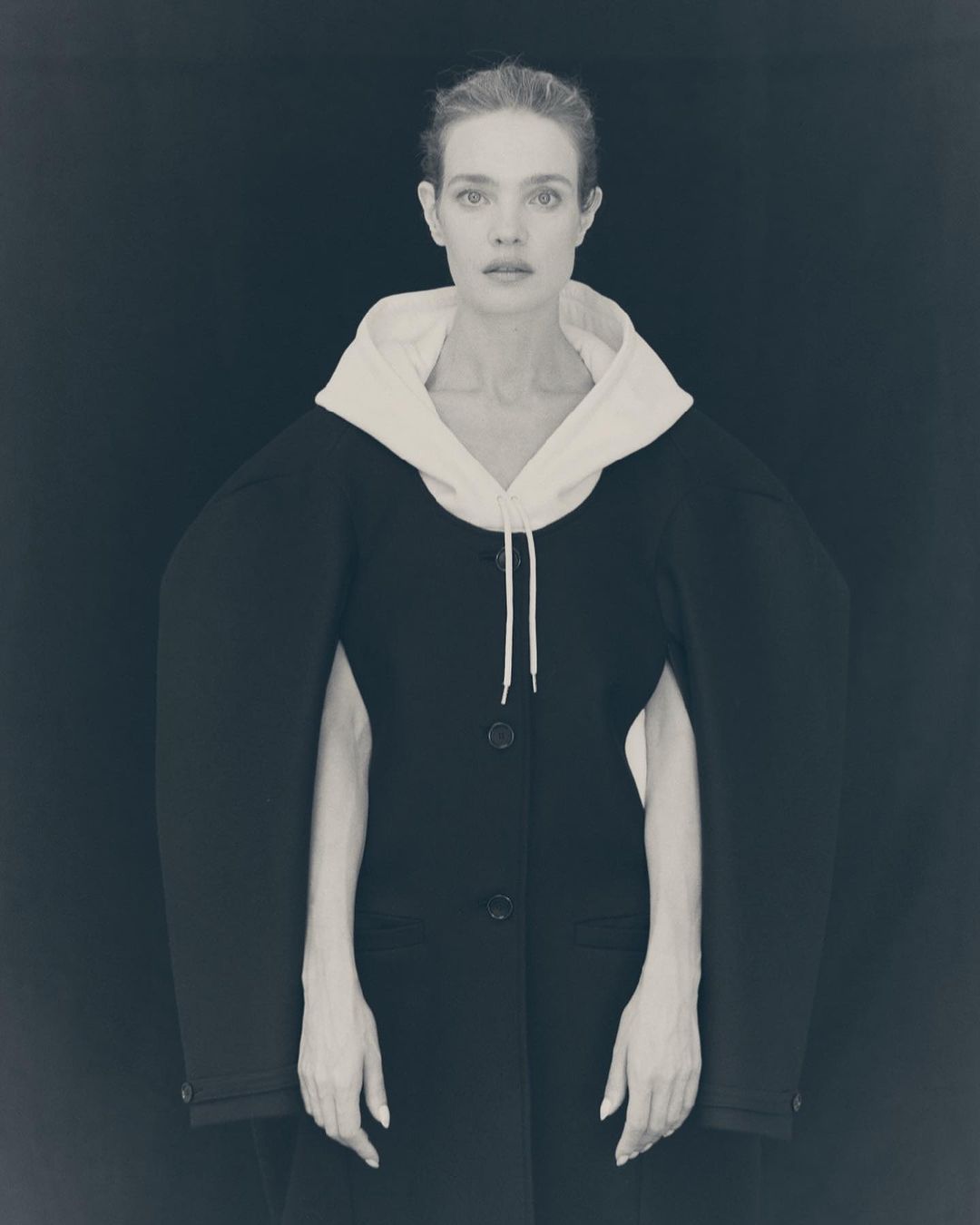 Monochrome portrait of a fair-skinned adult woman wearing a black coat with a white hood posing against a dark backdrop.