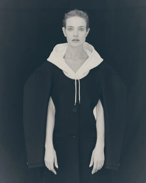 Monochrome portrait of a fair-skinned adult woman wearing a black coat with a white hood posing against a dark backdrop.