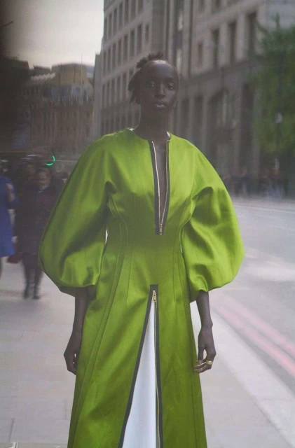 A fashion shot features an adult female model wearing a long, flowing green dress with puffed sleeves, standing on a city street.