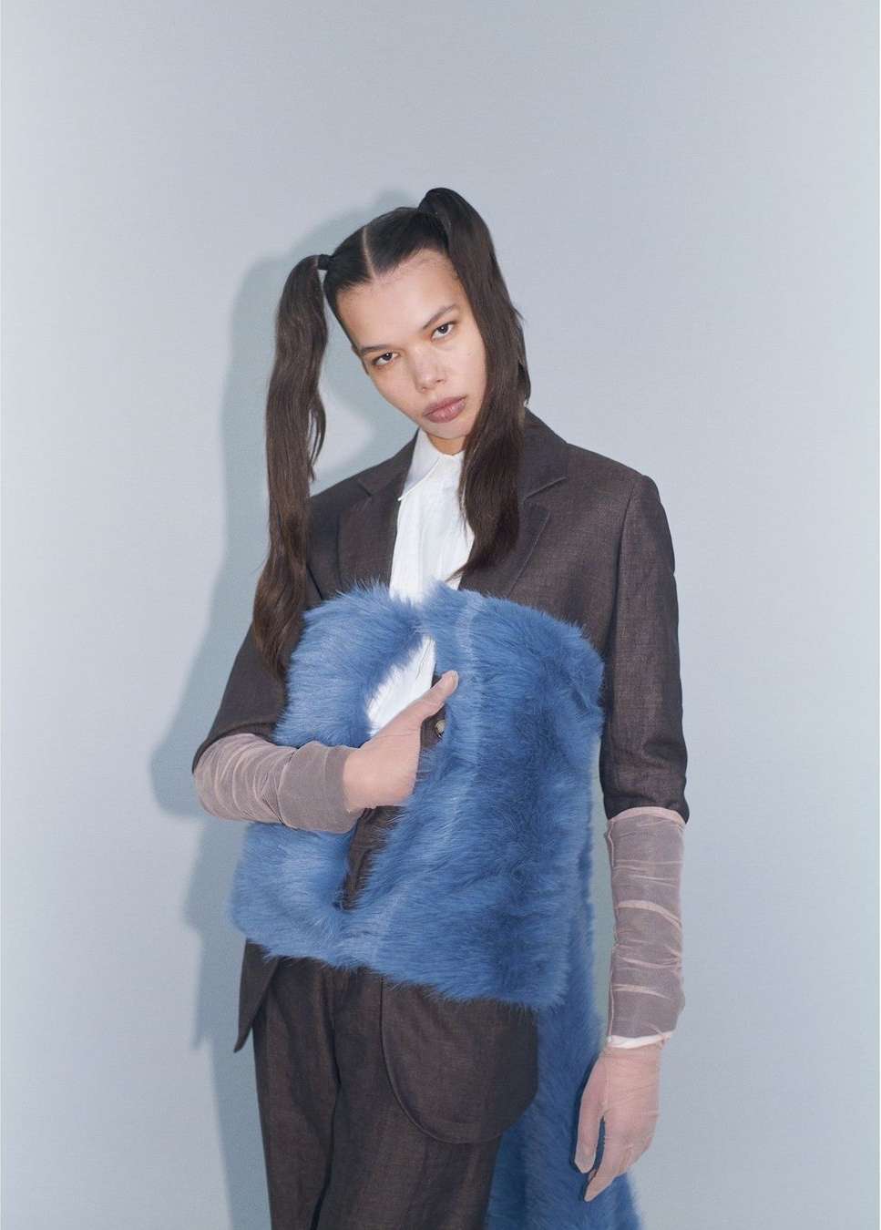 Fashionable young woman posing in a photo studio with a blue faux-fur bag, wearing a suit jacket and pigtails, against a neutral backdrop.