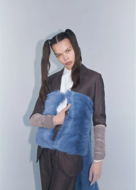 Fashionable young woman posing in a photo studio with a blue faux-fur bag, wearing a suit jacket and pigtails, against a neutral backdrop.