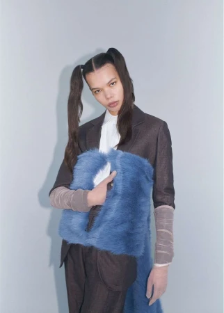 Fashionable young woman posing in a photo studio with a blue faux-fur bag, wearing a suit jacket and pigtails, against a neutral backdrop.