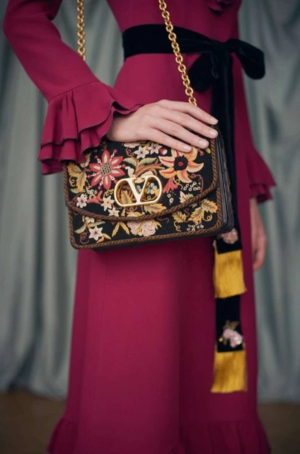 Close-up of a woman in a magenta dress holding a Valentino handbag with floral embroidery and gold hardware, displayed in a studio.