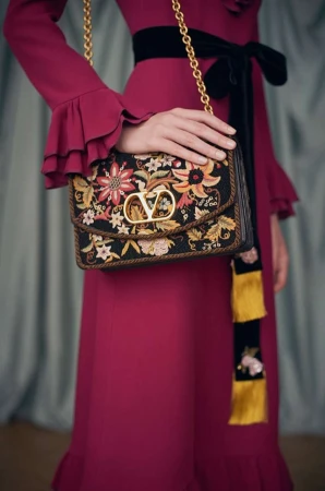 Close-up of a woman in a magenta dress holding a Valentino handbag with floral embroidery and gold hardware, displayed in a studio.