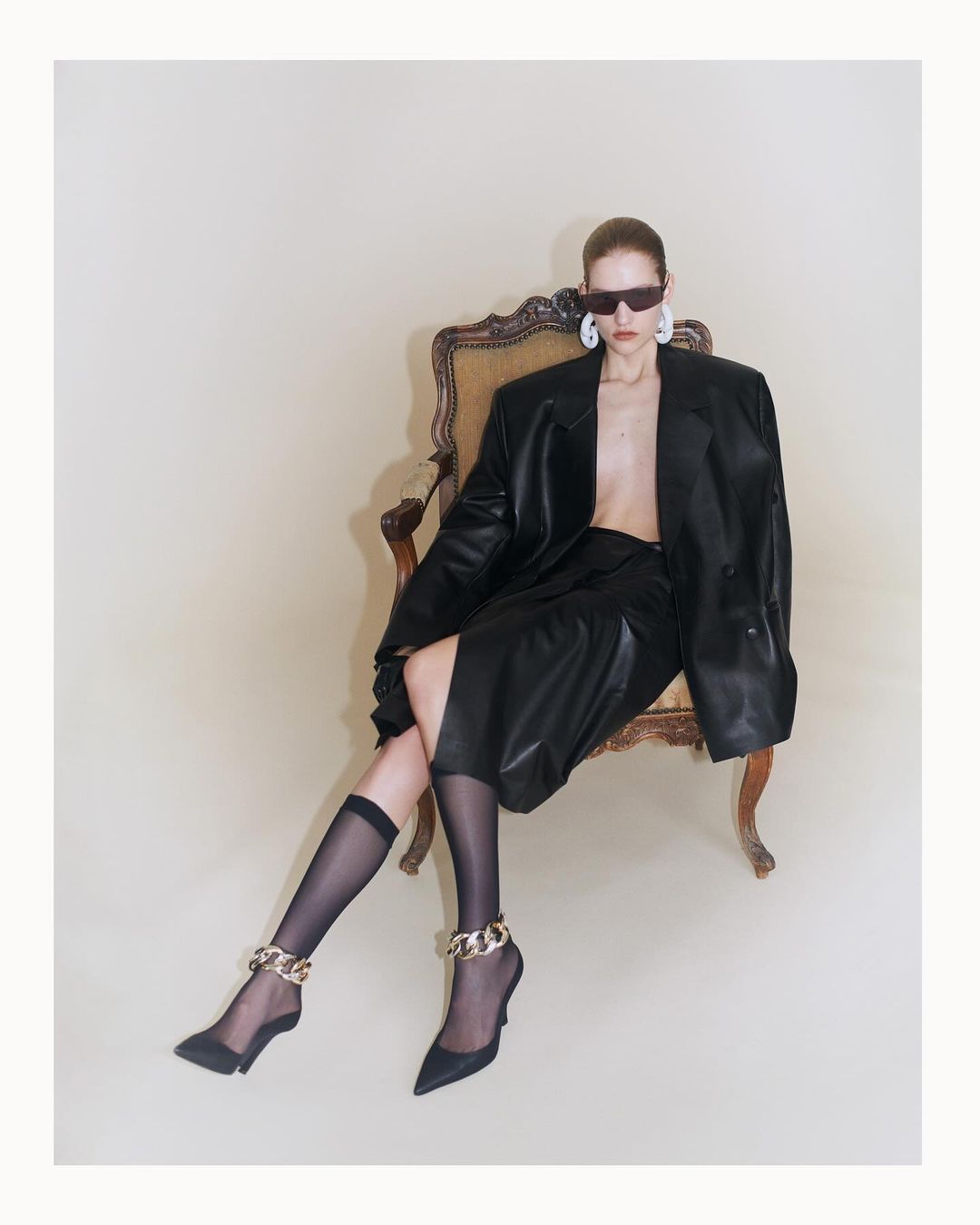 A model in sunglasses and an oversized black leather blazer poses on a chair with sheer socks and chain-strap heels against a plain background.