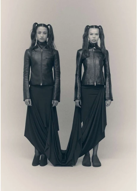 Two women with pigtails wearing matching black leather jackets and long black skirts pose in front of a neutral background.