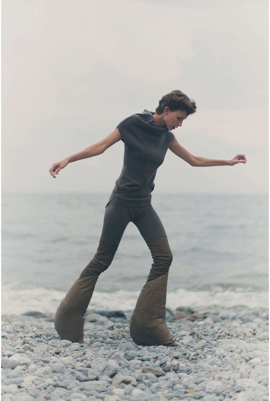 Woman on pebble beach wearing a gray outfit and large, padded shoes, walking by a gray ocean and overcast sky in an outdoor setting.